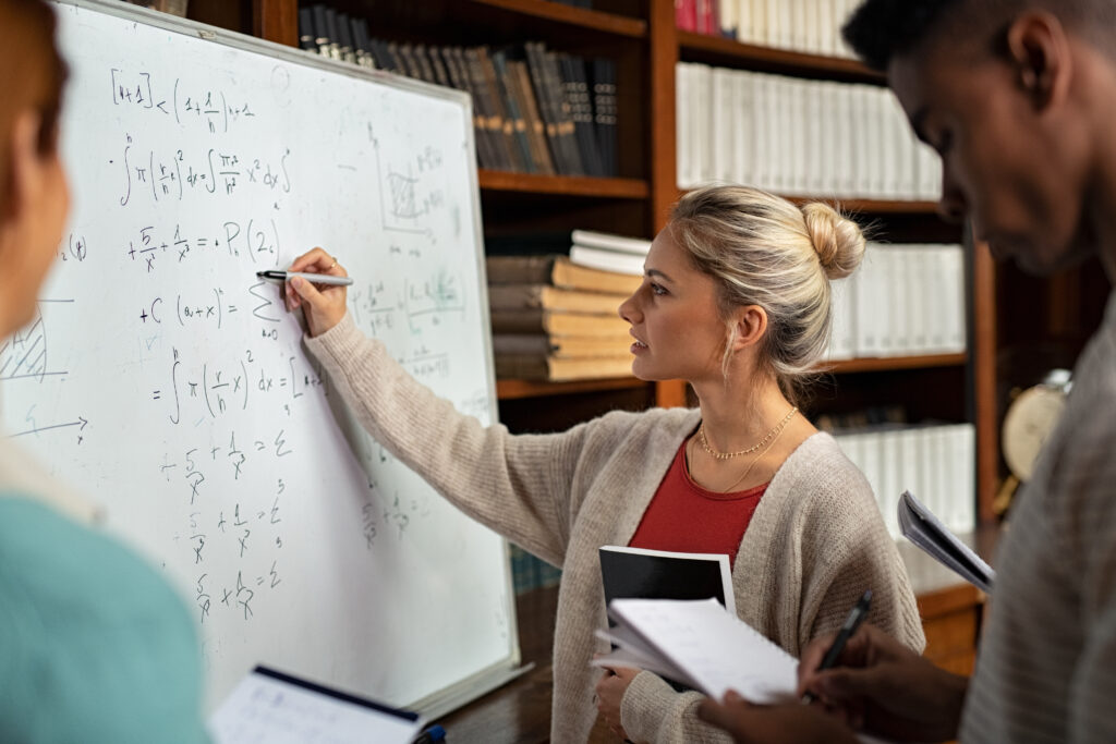 A young woman writes complex math equations on a whiteboard while two EWC students observe and take notes in a library or classroom setting filled with bookshelves.