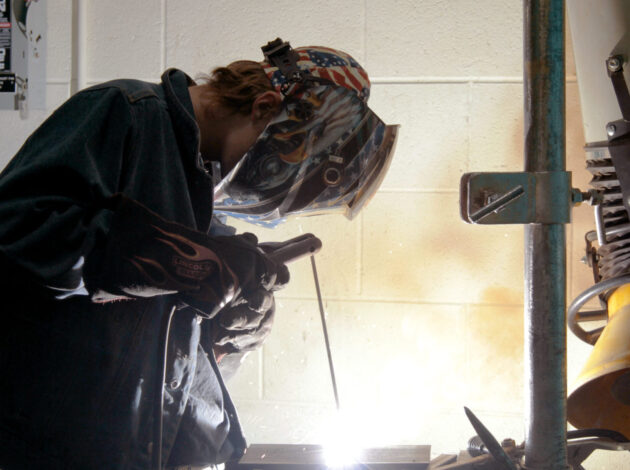 A person wearing an EWC-decorated welding helmet and gloves welds metal, with bright sparks and light visible near their hands in a workshop setting.