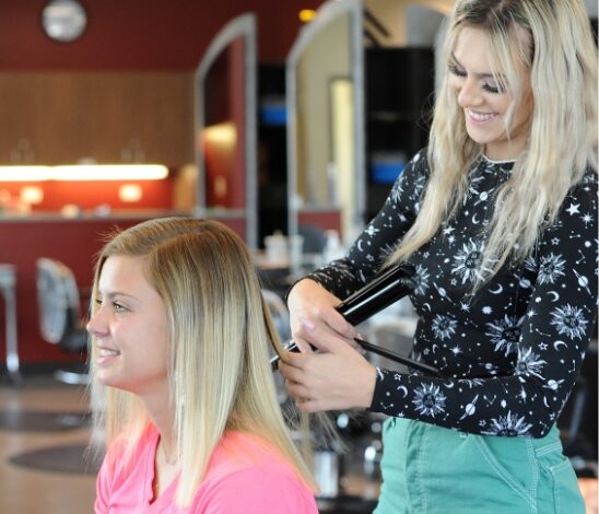 A hairstylist with long blonde hair, wearing green pants and a black patterned top, styles a smiling customer's straight blonde hair at an EWC salon. The customer sits in a chair wearing a pink shirt.