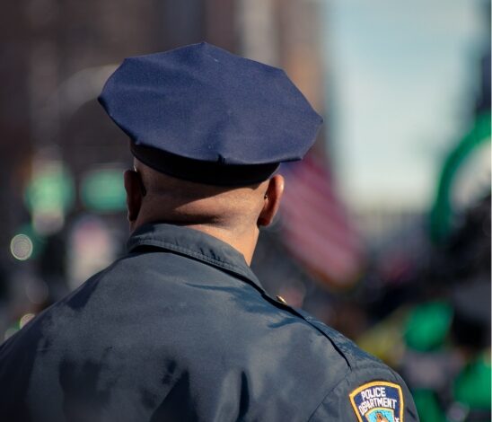 A police officer in uniform, seen from behind, stands outdoors with a blurred city background. The officer’s shoulder patch reads Police Department and features the EWC insignia.