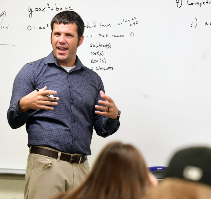 A man in a blue shirt stands in front of a whiteboard teaching an EWC class. The whiteboard has handwritten equations and a graph, while several students are seated and facing him, seen from behind.