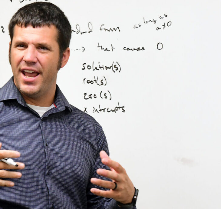 A man in a blue shirt stands in front of a whiteboard teaching an EWC class. The whiteboard has handwritten equations and a graph, while several students are seated and facing him, seen from behind.
