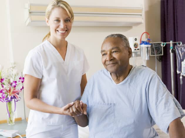 A nurse in white scrubs with an EWC badge smiles while holding hands with an older male patient sitting on a hospital bed. Medical equipment and a vase of flowers are visible in the background.