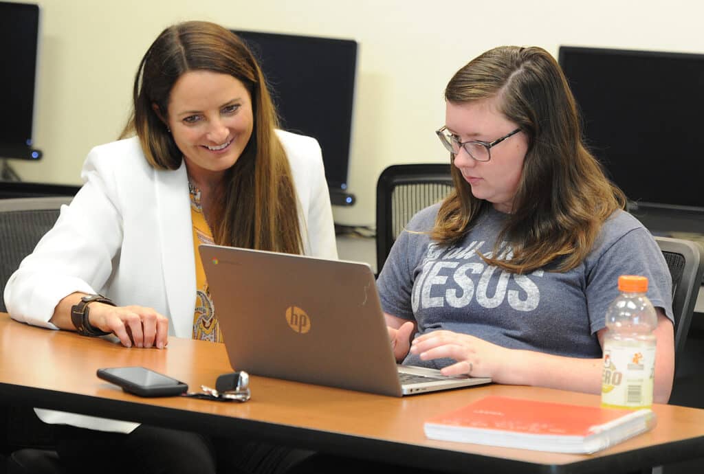 A woman in a white blazer smiles while sitting next to an EWC student typing on an HP laptop. The student wears glasses and a “Jesus” shirt. A phone, keys, notebook, and sports drink are on the table. Computers are in the background.