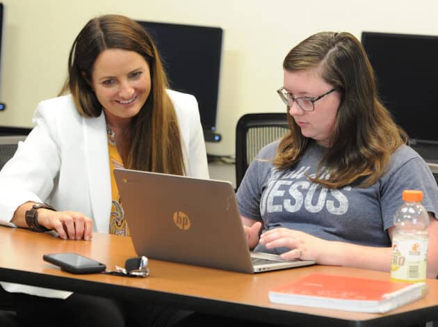 A woman in a white blazer smiles while sitting next to an EWC student typing on an HP laptop. The student wears glasses and a “Jesus” shirt. A phone, keys, notebook, and sports drink are on the table. Computers are in the background.