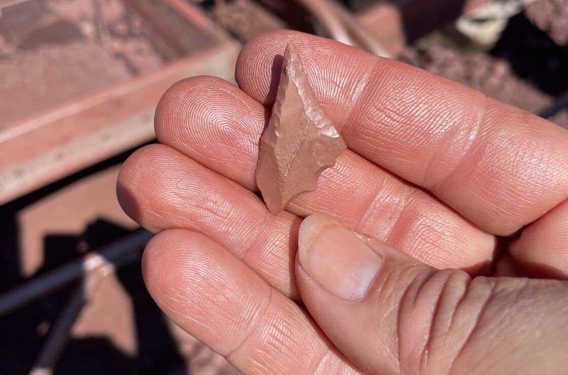 A hand holds a small, pointed stone tool or artifact with reddish-brown coloration, possibly uncovered during an EWC excavation. Blurred excavation equipment and soil are visible in the background.