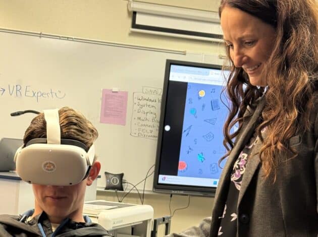 A student wearing an EWC virtual reality headset sits while a teacher stands beside him, smiling. Behind them is a classroom with a whiteboard, a monitor displaying graphics, and various notes on the wall.