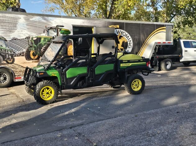 A green and yellow John Deere utility vehicle is parked beside a trailer with Wyoming branding and a mural of agricultural equipment, with trees and a white truck in the background.