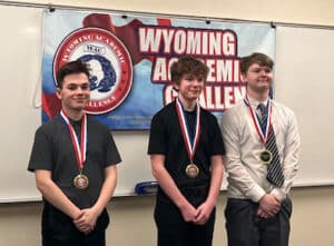 Three teenage boys wearing medals stand smiling in front of a Wyoming Academic Challenge banner in a classroom setting.