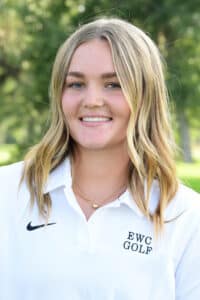 A young woman with long blonde hair smiles outdoors, wearing a white collared shirt with EWC GOLF and a Nike logo, standing in front of green trees.