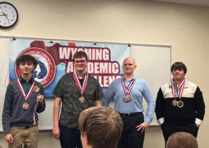 Four people stand in front of a Wyoming Academic Challenge banner, wearing medals around their necks. One person holds up a medal. An audience members head is visible in the foreground. A clock is on the wall.