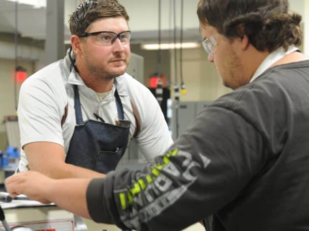 Two men wearing safety glasses and work clothes have a serious conversation in an industrial workshop, surrounded by equipment and machinery. welding joel