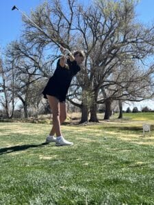 A person swings a golf club on a grassy course with tall trees in the background under a clear blue sky. They are mid-swing, wearing a dark shirt, shorts, and white athletic shoes.