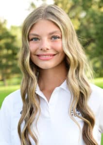 A young woman with long, wavy blonde hair and blue eyes smiles while standing outdoors. She is wearing a white collared shirt and is in front of a background of green trees.