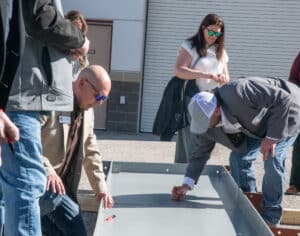 A group of people stands outdoors near a metal structure; two men kneel and write on the structure while others watch. One woman stands in the background with her coat draped over her arm.