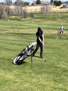 A person wearing black pants and a black hat is swinging a golf club on a grassy course, with a golf bag nearby. Another golfer is visible in the background under a clear sky.