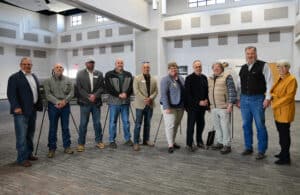 Ten adults stand in a row inside a spacious, well-lit room with gray carpet and white walls. Most wear business casual or western attire, and they are smiling at the camera.