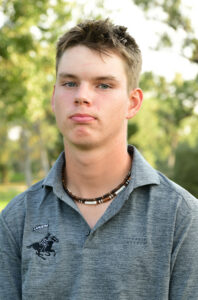 A young man with short brown hair wears a gray polo shirt with a Lancers logo and a beaded necklace, standing outdoors with green trees blurred in the background. He has a neutral expression.