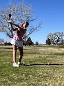 A young woman in a pink tank top and white skirt prepares to swing a golf club on a sunny day, standing on a grassy golf course with trees and a clear blue sky in the background.