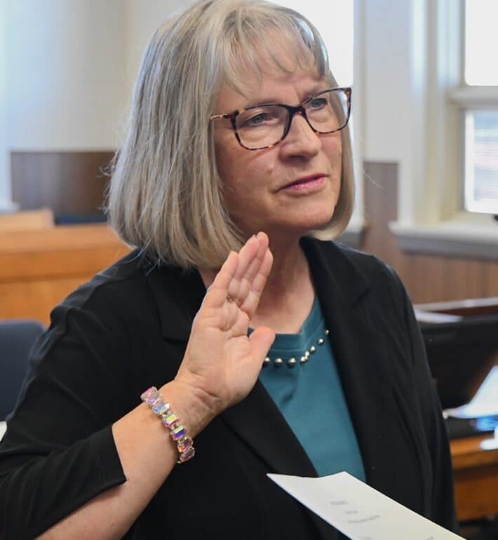 A woman with gray hair and glasses raises her right hand while holding a document in her left, appearing to take an oath in a courtroom setting. She is wearing a teal top, black jacket, and a beaded bracelet.