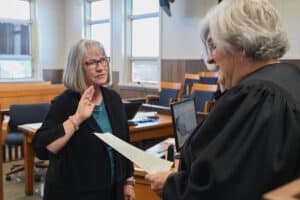 A woman stands with her right hand raised, being sworn in by a judge holding a document in a courtroom. Both women have gray hair and wear glasses. The background shows empty chairs and large windows.