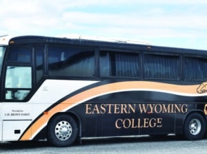 A black bus with orange and white accents displays Eastern Wyoming College on its side. The bus is parked outdoors on a gravel surface under a cloudy sky.