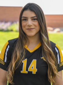 A young woman with long, brown, highlighted hair smiles at the camera, wearing a black and gold athletic jersey with the number 14 printed on the front, set against a blurred green and brick outdoor background.