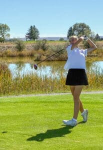 A young woman wearing a white shirt and black skirt swings a golf club on a grassy course near a pond, with trees and blue sky in the background.