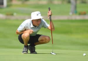 A golfer wearing a white hat and polo squats on the green, closely eyeing a putt with a putter in hand. A golf ball rests near the hole on a sunny day.