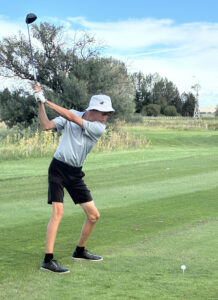 A person wearing a light bucket hat, gray shirt, and black shorts is poised to hit a golf ball on a green course, with trees and a windmill in the background.