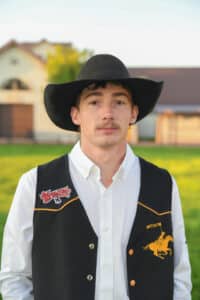 A young man in a black cowboy hat and vest with a white shirt stands outside on grass, with buildings blurred in the background.