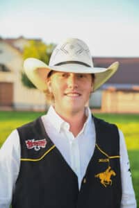 A young person in a white cowboy hat and vest stands outside, smiling. The vest features a horse logo and a Wyoming patch. There are buildings and grass in the blurred background.