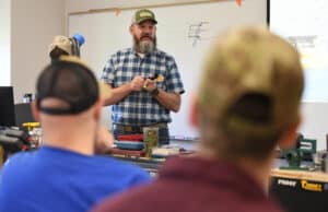 Bearded man in a plaid shirt and cap speaks to a classroom, holding a small object, while two people in caps listen in the foreground. Workshop tools and diagrams are visible in the background.