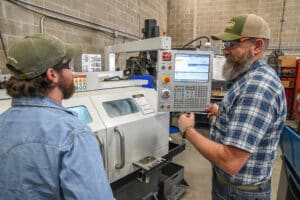 Two men wearing safety glasses and hats operate a CNC machine in an industrial workshop. One man points at the control panel screen while explaining the process to the other.