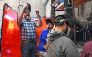 Three people in welding helmets work in an industrial workshop. One man in a plaid shirt stands adjusting his helmet, while two others sit and prepare to weld. Red safety screen and equipment are visible in the background.