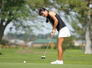 A woman wearing a black sleeveless top and white skirt is putting on a golf course, focusing on the ball. Trees and greenery are visible in the background.