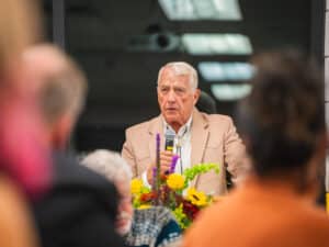 An older man in a tan blazer speaks into a microphone at a podium decorated with flowers, viewed from behind two people in the foreground.