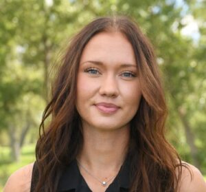 A young woman with long wavy brown hair wearing a black sleeveless polo shirt and a small pendant necklace.