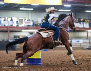A cowgirl in a cowboy hat and vest riding a horse around a blue and yellow barrel during a barrel racing competition.